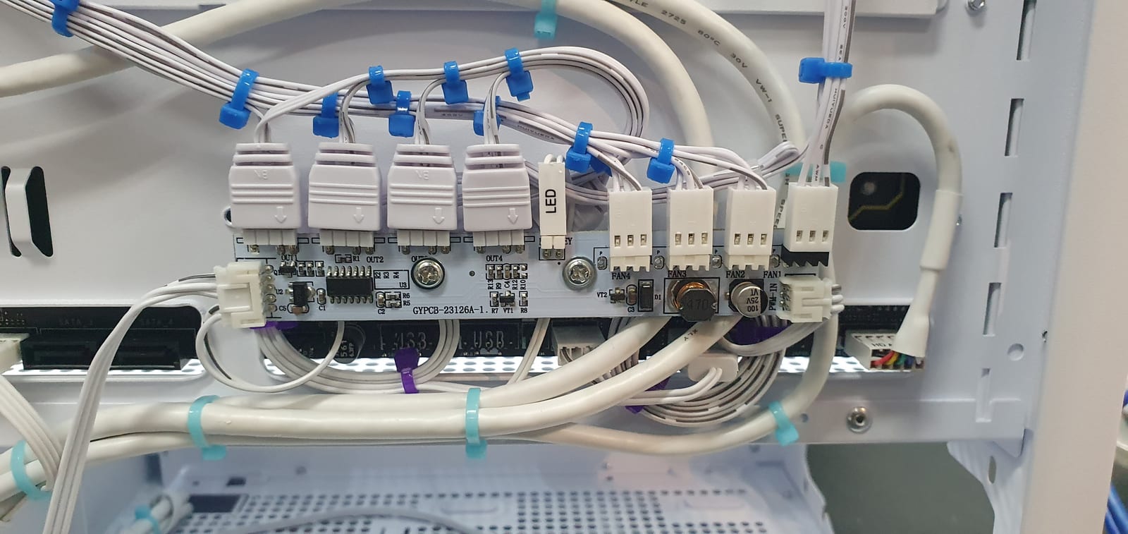 Behind the motherboard tray of a white PC case, where fan and lighting cables are routed to a hub and held with blue and turquoise clips grouped by function