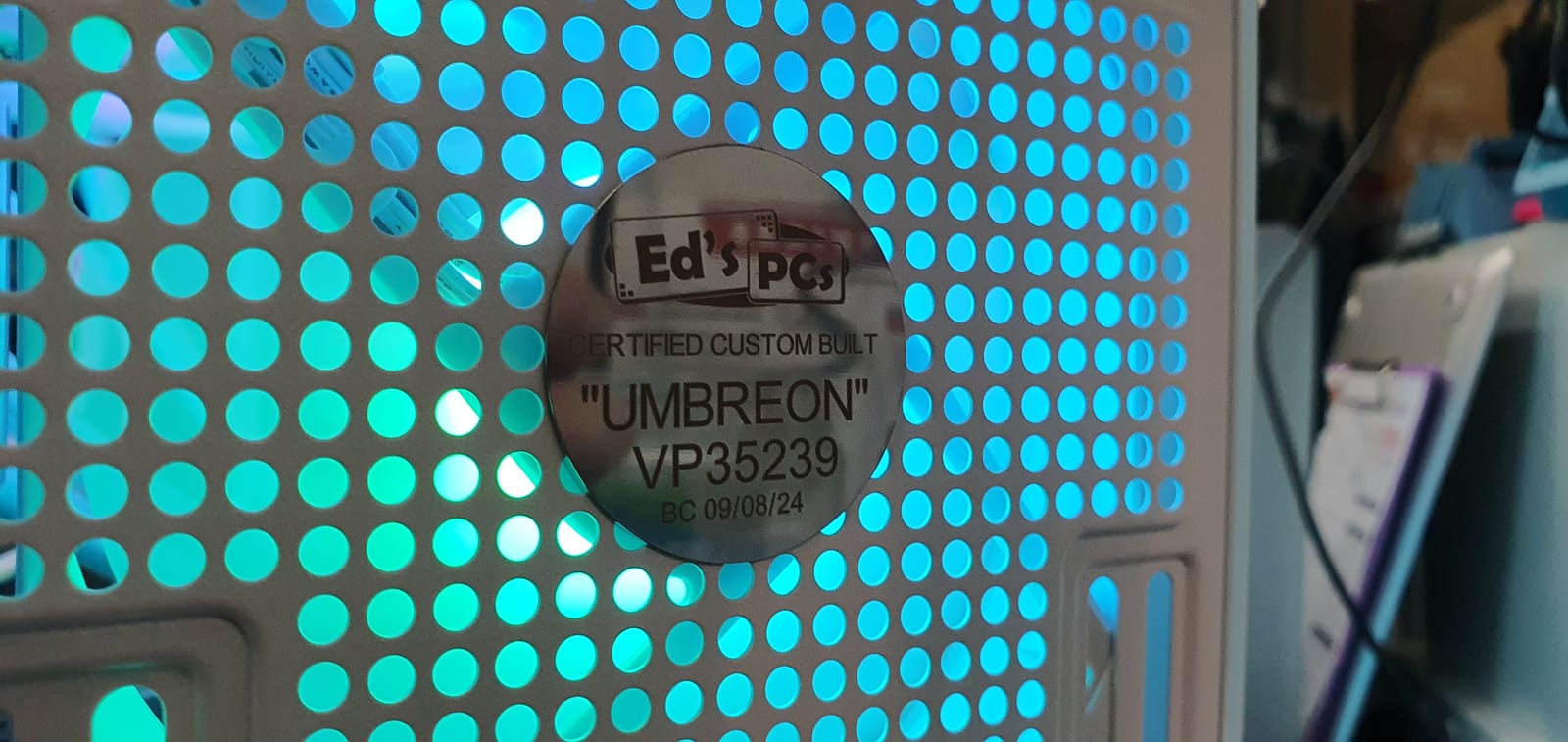 A round engraved metal plate reading Ed's PCs, Certified Custom Built, "UMBREON", fixed to a perforated case panel backlit in cyan