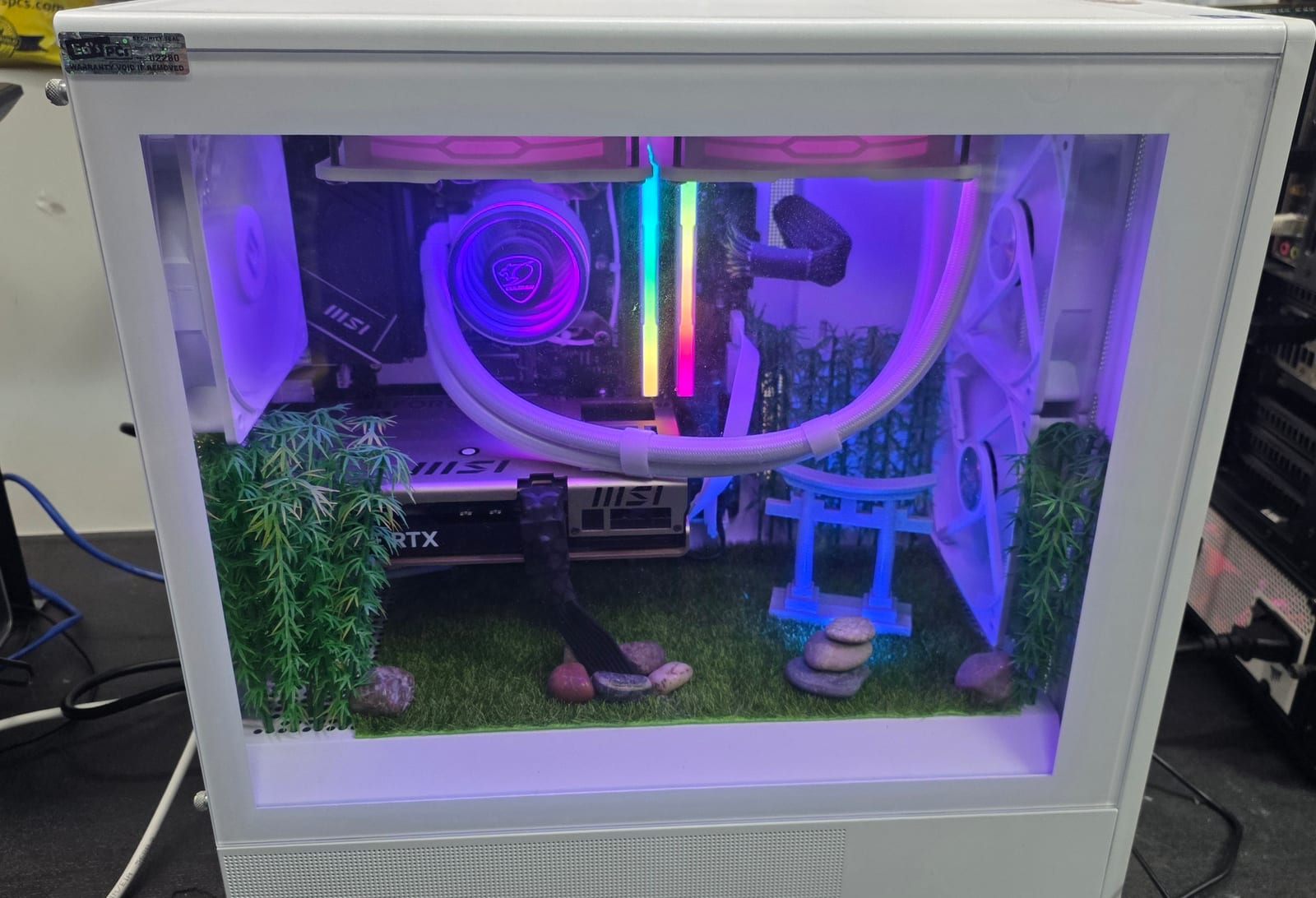 A white PC case with a Japanese garden diorama in the lower chamber — turf, bamboo, a torii gate and stacked stones — under a white-sleeved cooling loop lit violet