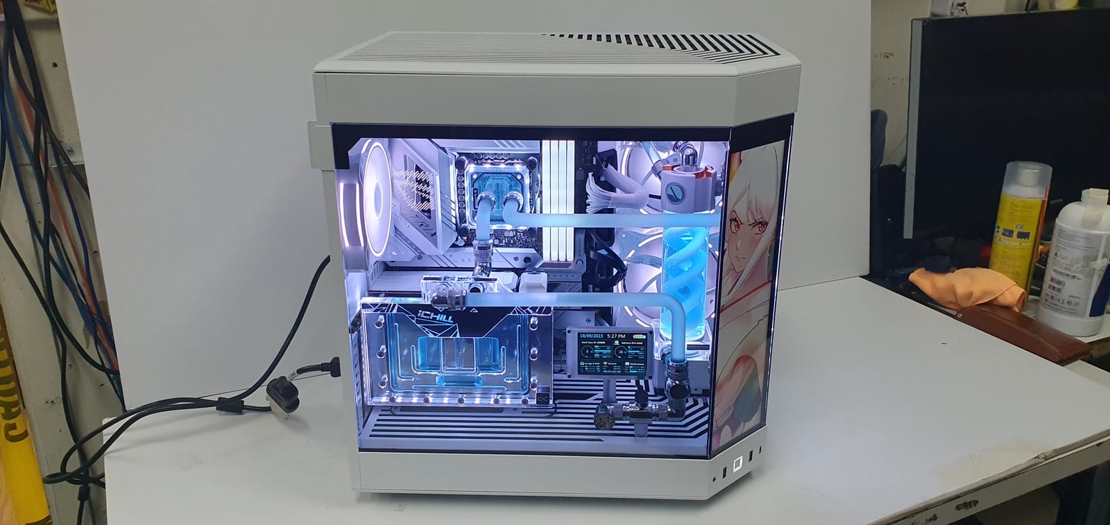A white dual-chamber PC case on a workbench with a lit blue hard-line water cooling loop, distribution plate and a tall display panel behind the front glass