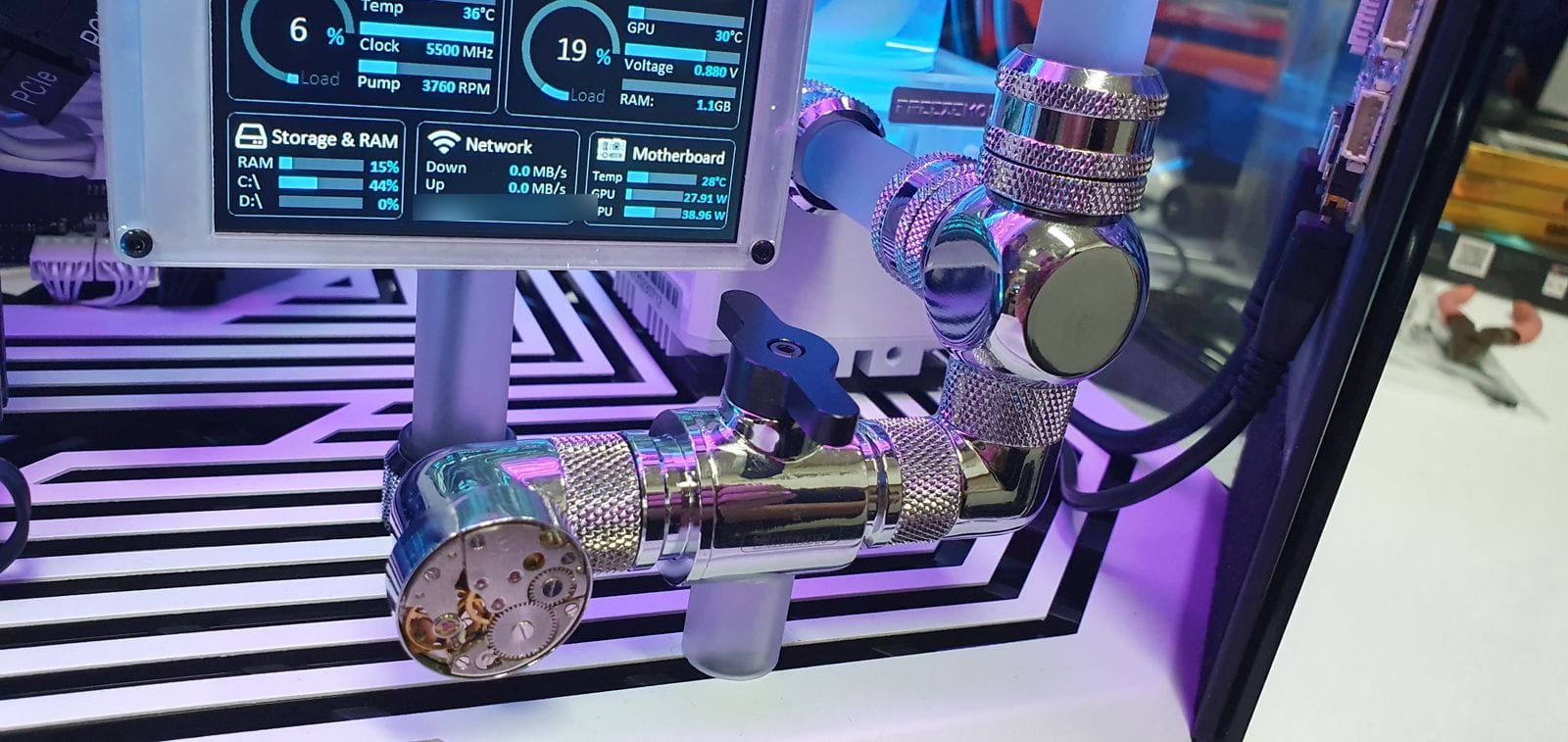 A close view of the in-case sensor panel showing processor load, clock speed, pump RPM, graphics temperature, storage, network and motherboard power draw, beside chrome fittings and a watch movement