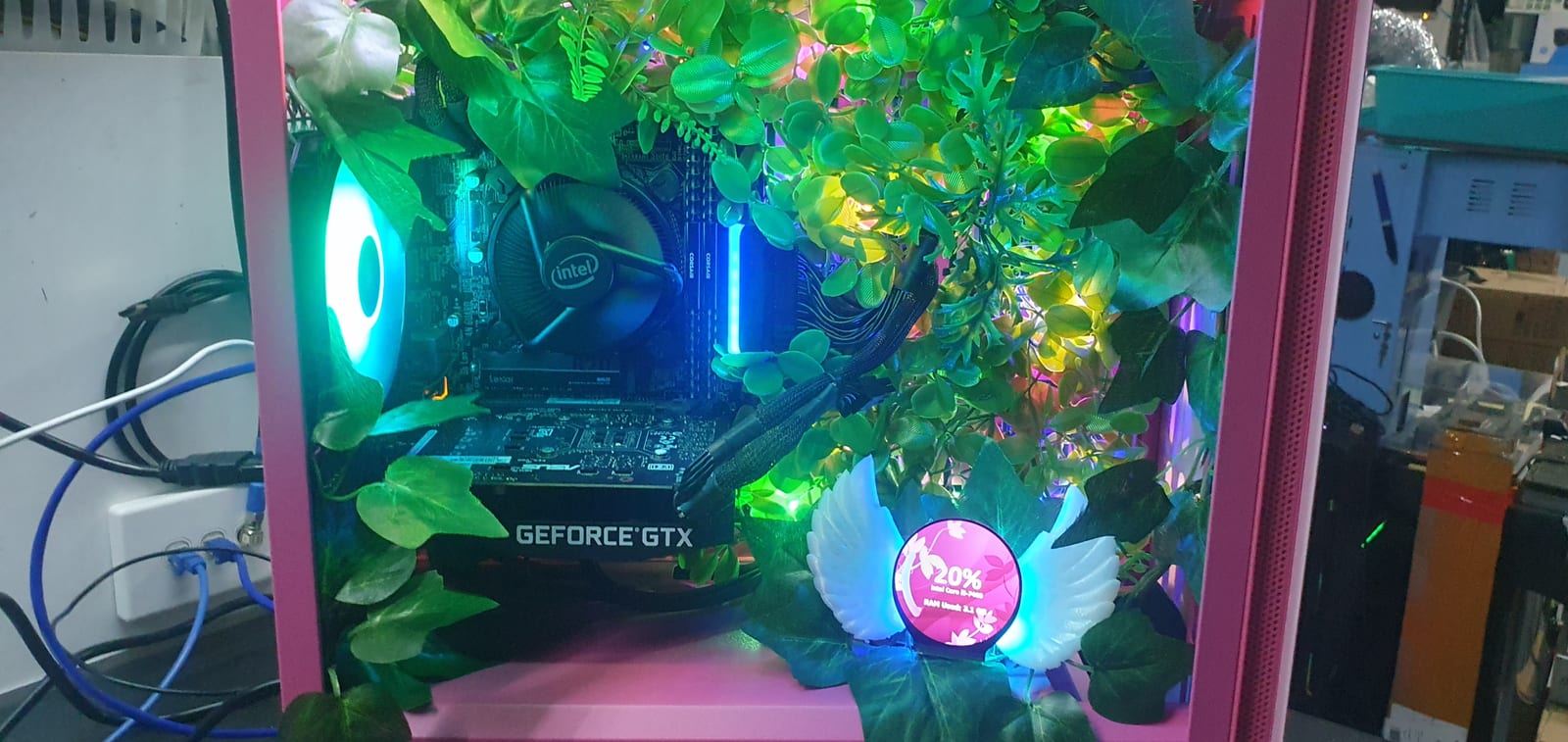 A pink PC case with green foliage arranged around the motherboard and graphics card, and a round winged display showing processor and memory usage
