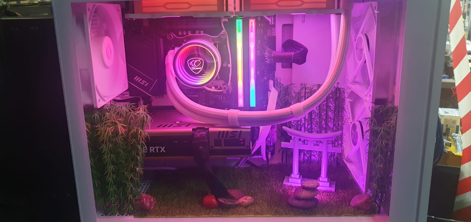 A white PC case lit pink and purple, with a miniature Japanese garden of turf, bamboo and a torii gate built into the floor beneath the components