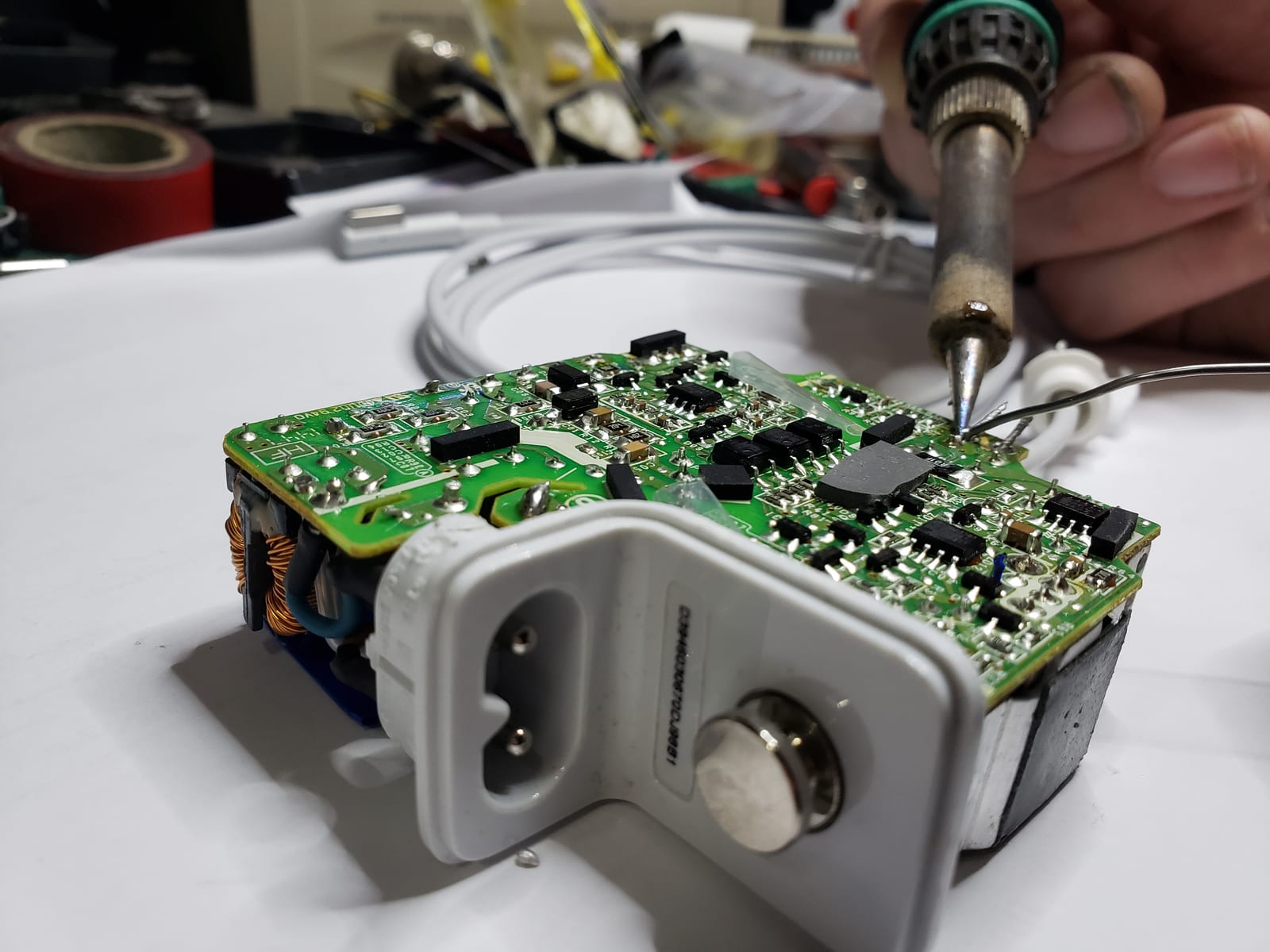 A hand holding a soldering iron and solder wire over the opened circuit board of a laptop power adapter on the workbench
