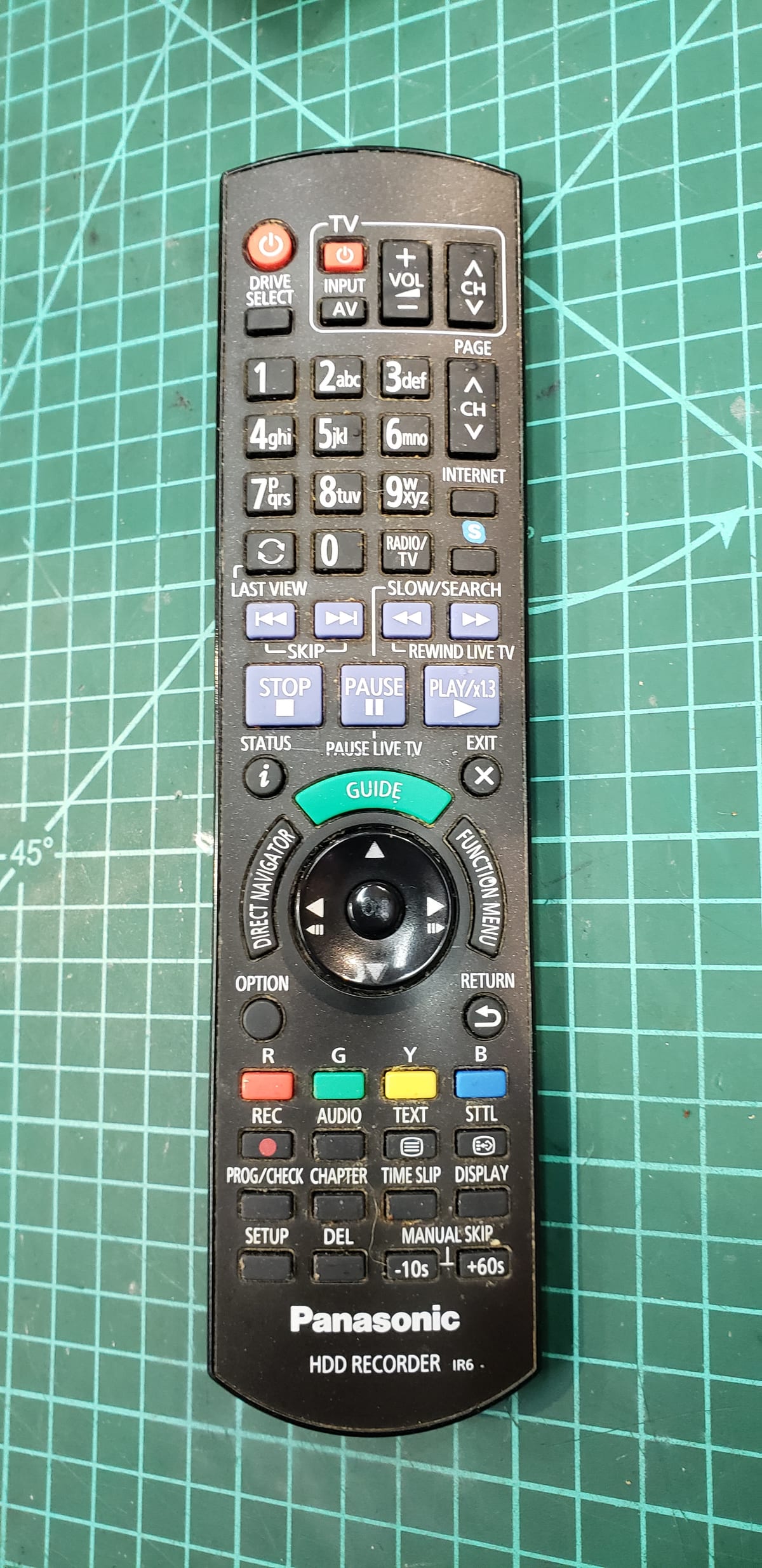 A grimy black Panasonic HDD recorder remote control on a green cutting mat, buttons discoloured with years of use